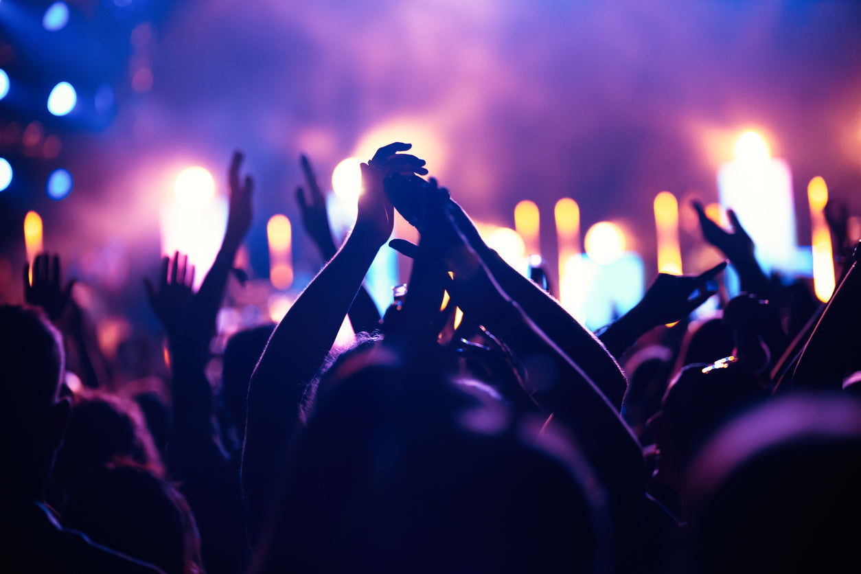 Crowd at a live concert with raised hands silhouetted against colorful stage lights.